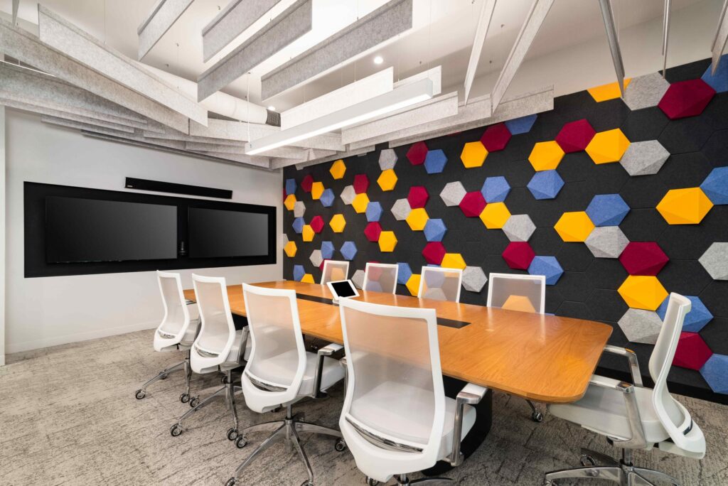 A vibrant and modern conference room with a long wooden table, white office chairs, and two large mounted screens. The room features a striking accent wall with colorful hexagonal acoustic panels in red, blue, yellow, and gray, and the ceiling is decorated with gray and white acoustic panels.