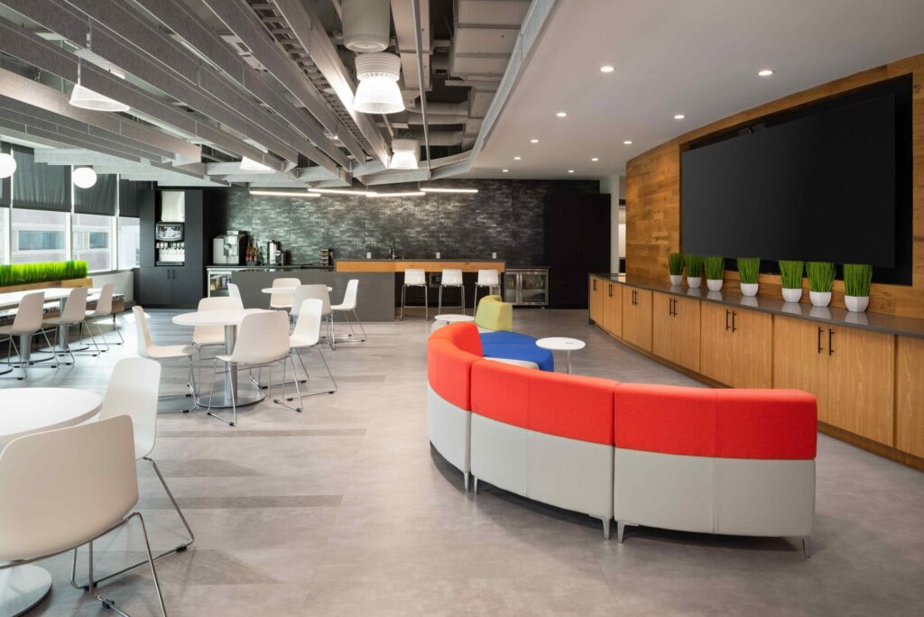 An open-plan office breakroom with a large curved sofa, tables and chairs, a kitchen counter, and a large wall-mounted television.